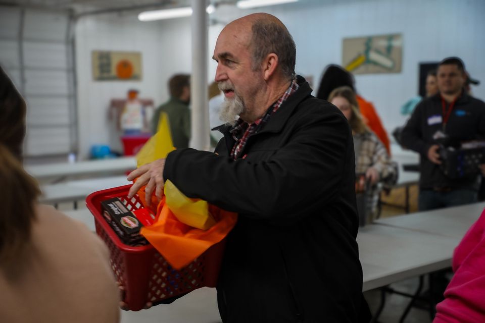 A volunteer with a white beard carries a red basket of Thanksgiving groceries tucked with orange and yellow tissue paper.