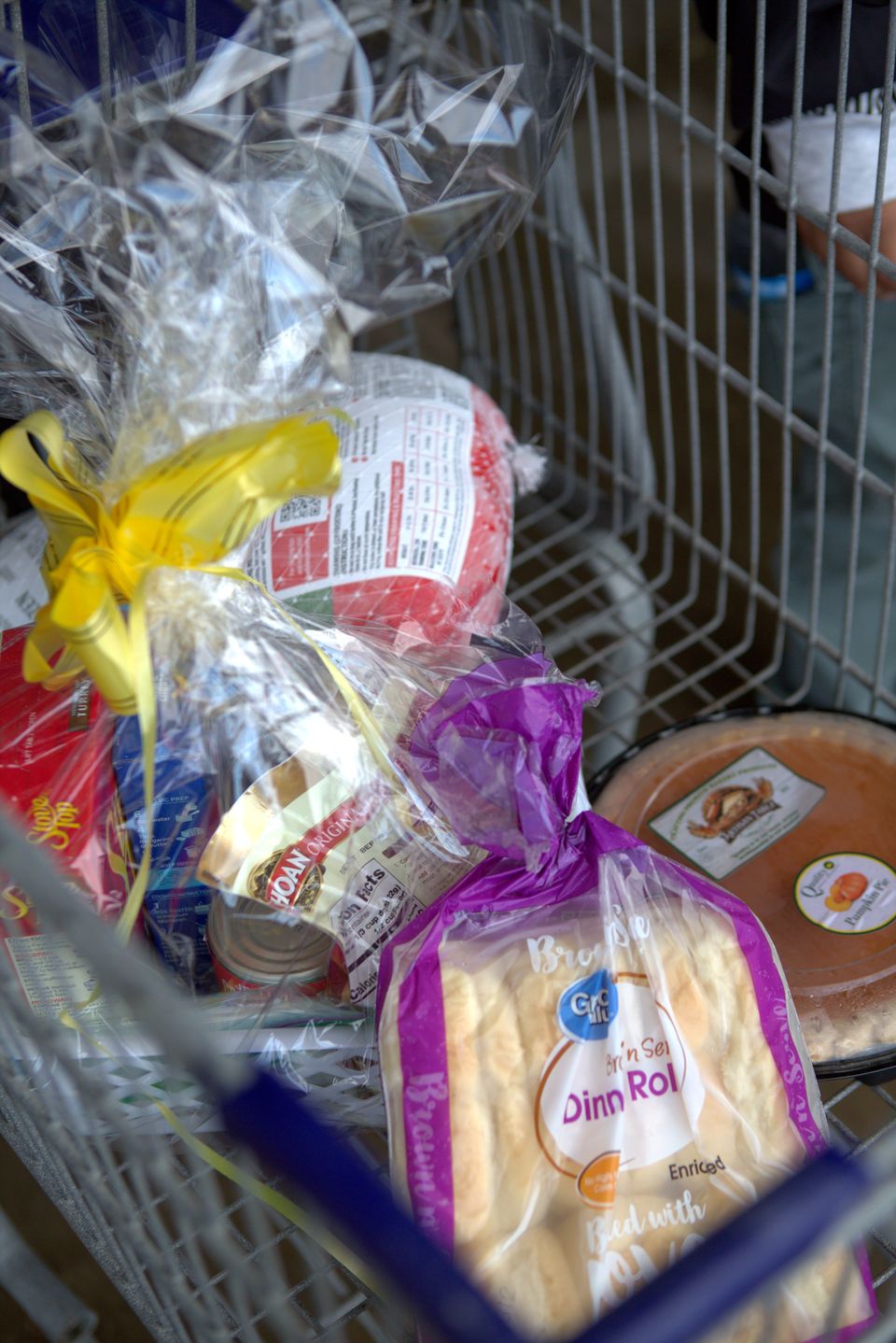 A finished basket in a cart: a turkey, stuffing, canned vegetables, dinner rolls, and a pumpkin pie, wrapped in cellophane and yellow ribbon.
