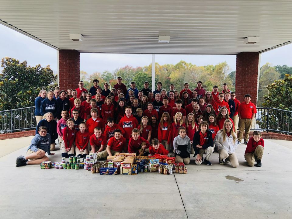 A school group in red and navy shirts gathers under a pavilion behind rows of donated Thanksgiving food.