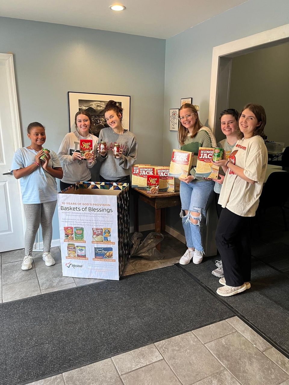 Six young volunteers hold up boxes of mashed potatoes, stuffing, and canned green beans around a Baskets of Blessings collection box.