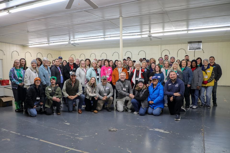 About sixty Baskets of Blessings volunteers with name tags gather for a group photo in the packing hall.