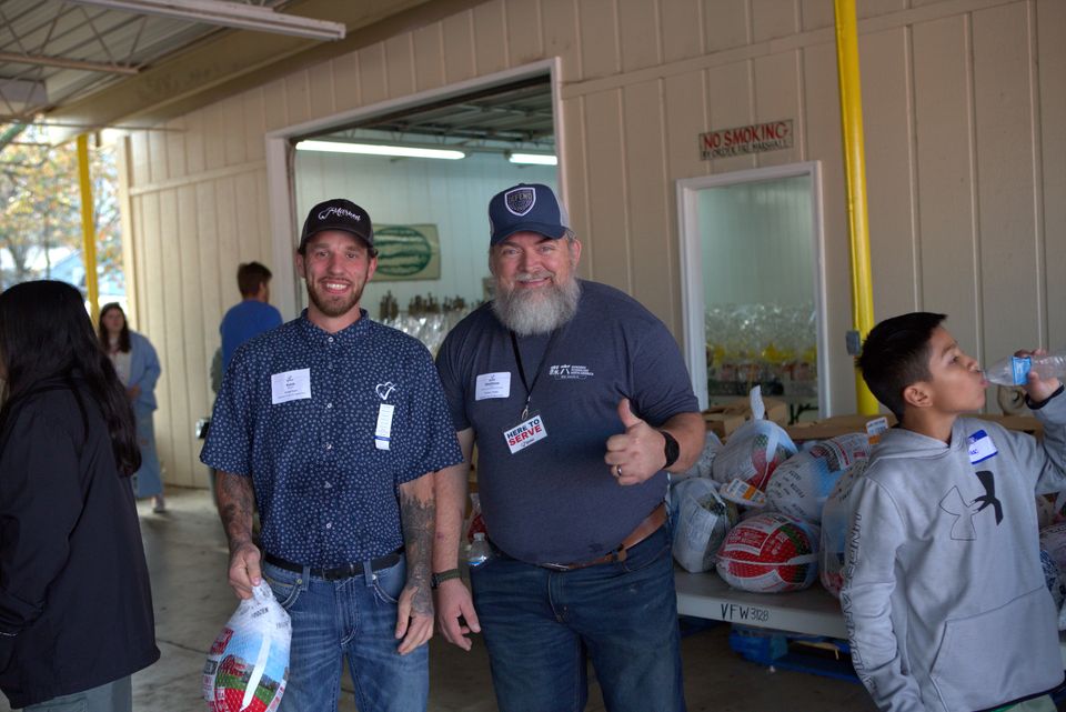 Two volunteers with Here to Serve badges smile at Distribution Day, one holding a frozen turkey, beside a table stacked with turkeys.