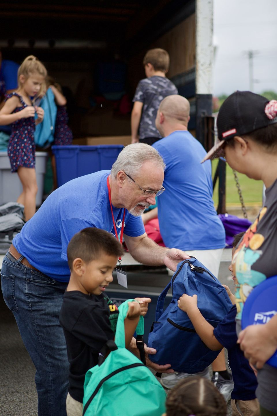 A volunteer in a blue Marked For Life shirt hands a navy backpack to a boy at the back of a delivery truck during Back to School Outreach.
