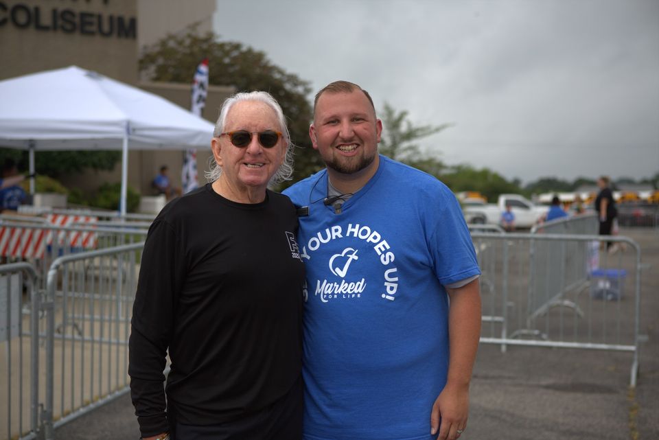 Logan LeGrande in a blue Get Your Hopes Up shirt stands beside an older volunteer outside the coliseum at a Deliver Hope event.