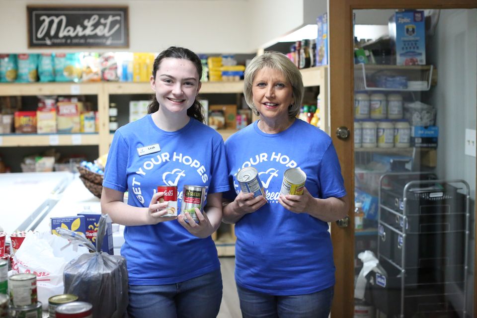 Leah and a fellow volunteer in blue Get Your Hopes Up shirts hold up canned goods in the Hope Center Market.