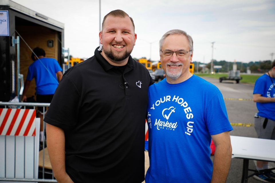 Logan LeGrande and a volunteer in a blue Get Your Hopes Up shirt smile beside the Deliver Hope trailer at an outreach event.