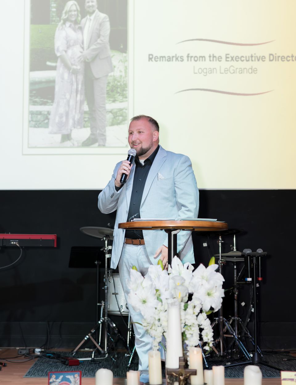 Logan LeGrande speaks at a Hope House graduation beneath a slide reading Remarks from the Executive Director.