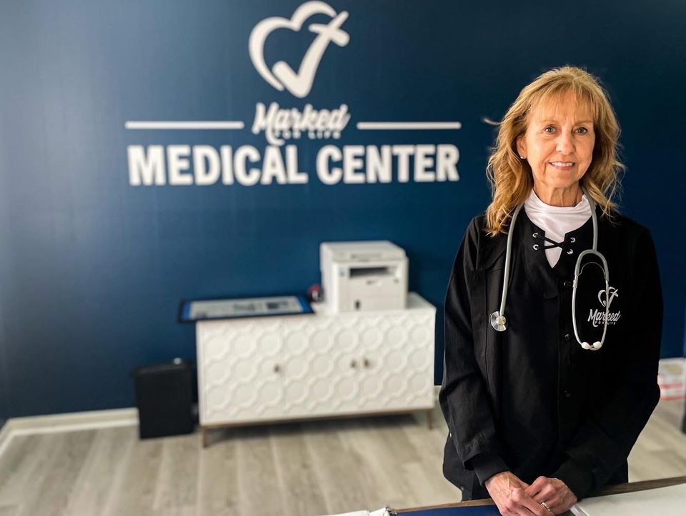 A nurse with a stethoscope stands at the front desk beneath the Marked For Life Medical Center wall sign.